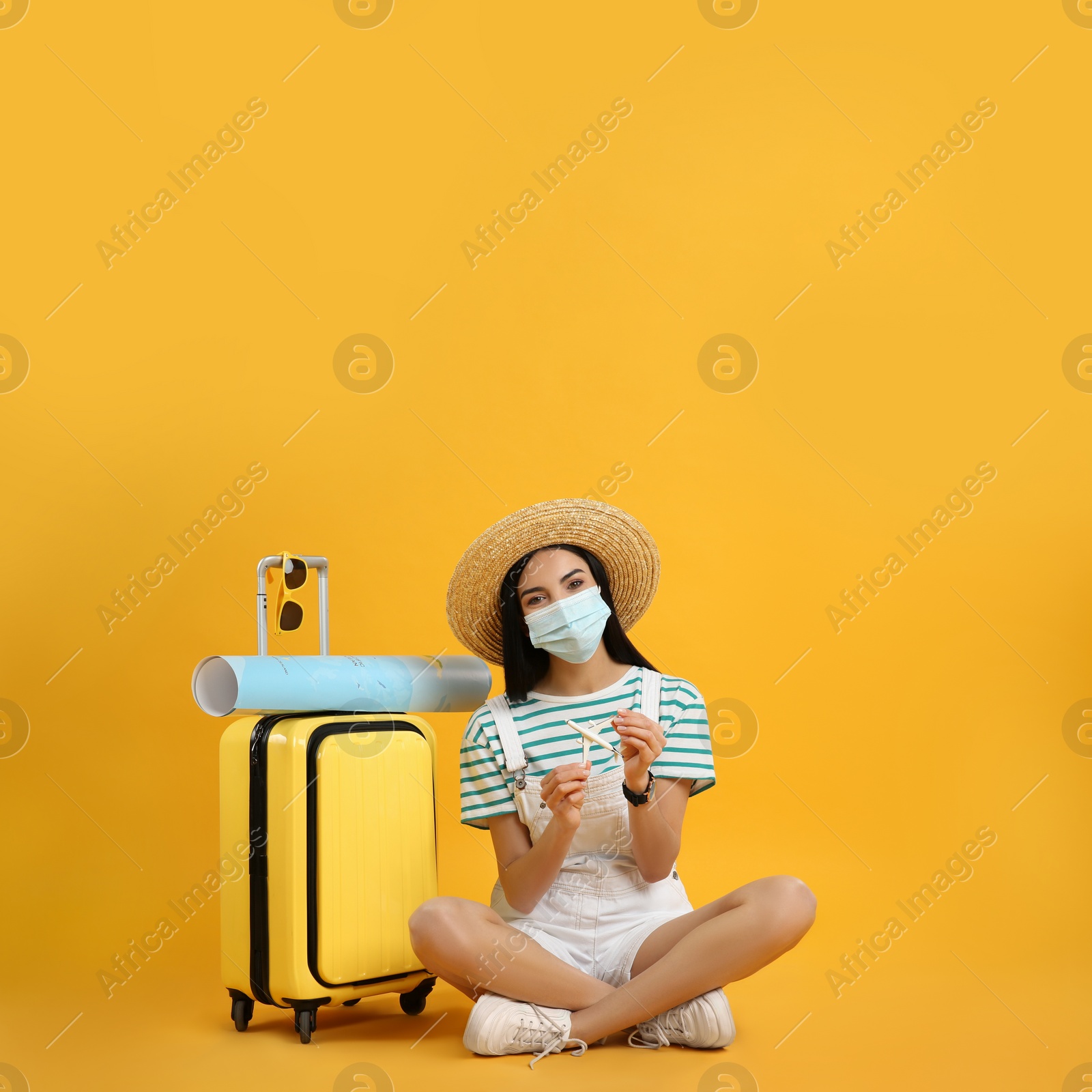 Photo of Female tourist in medical mask with suitcase, map and toy plane on yellow background. Travelling during coronavirus pandemic