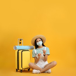 Photo of Female tourist in medical mask with suitcase, map and toy plane on yellow background. Travelling during coronavirus pandemic