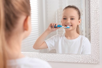 Photo of Cute little girl brushing her teeth with plastic toothbrush near mirror indoors