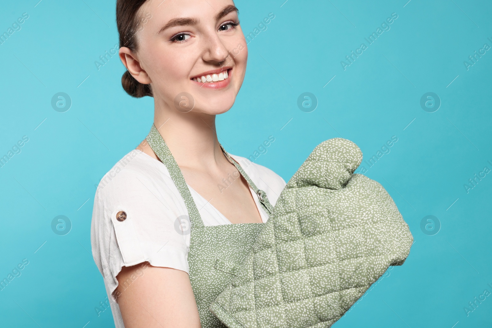 Photo of Beautiful young woman in clean apron with pattern and oven glove on light blue background, closeup