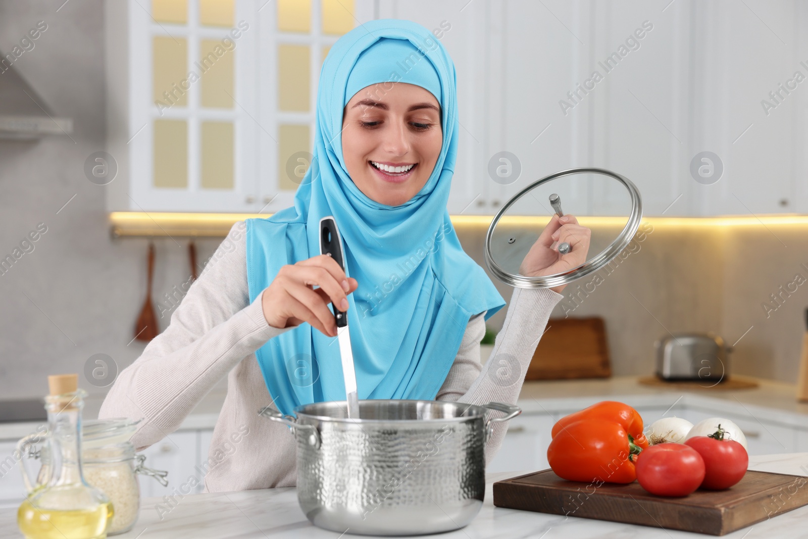 Photo of Muslim woman making delicious soup with vegetables at white table in kitchen
