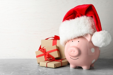 Photo of Piggy bank with Santa hat and gift boxes on grey table. Space for text