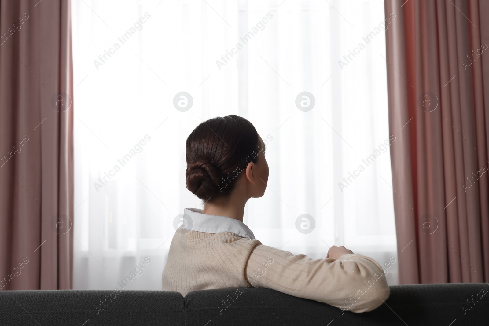 Photo of Woman sitting on sofa near window with stylish curtains at home, back view