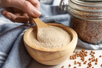 Woman taking buckwheat flour with spoon from bowl at white table, closeup