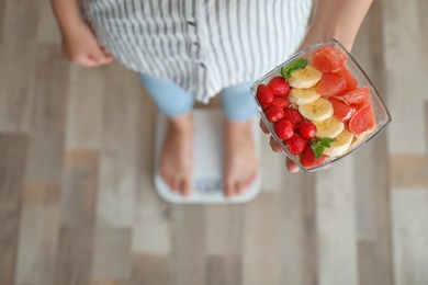 Photo of Woman holding bowl with delicious organic meal while measuring her weight on floor scales, top view. Healthy diet