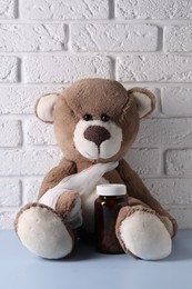 Photo of Toy bear with bandage and bottle of pills on light blue table near white brick wall