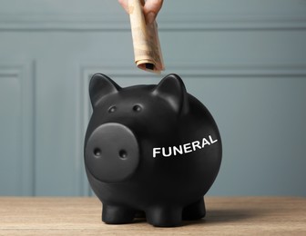 Money for funeral expenses. Woman putting banknotes into black piggy bank at wooden table, closeup