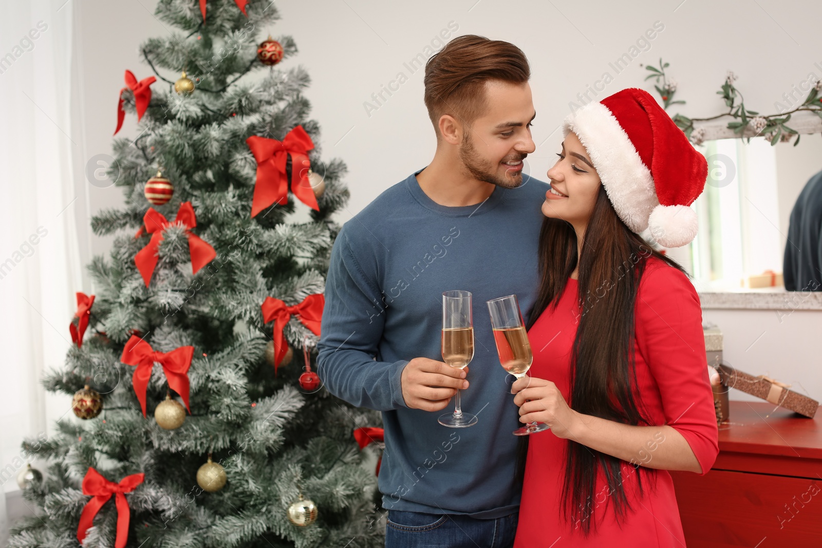 Photo of Young beautiful couple with glasses of champagne near Christmas tree at home