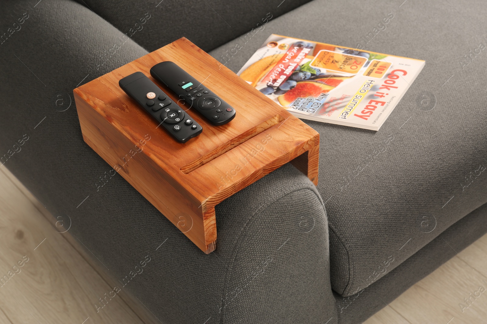 Photo of Magazine and remote controls on sofa with wooden armrest table in room. Interior element