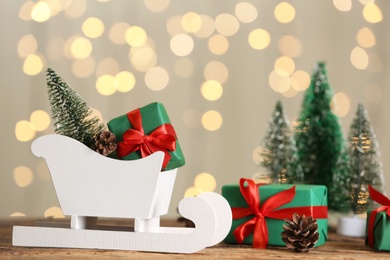 Photo of Small white sleigh, decorative Christmas trees and gift boxes on wooden table against blurred lights