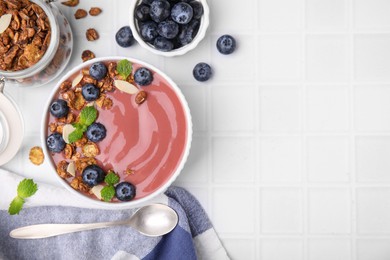 Photo of Bowl of delicious smoothie served with fresh blueberries and granola on white tiled table, flat lay. Space for text