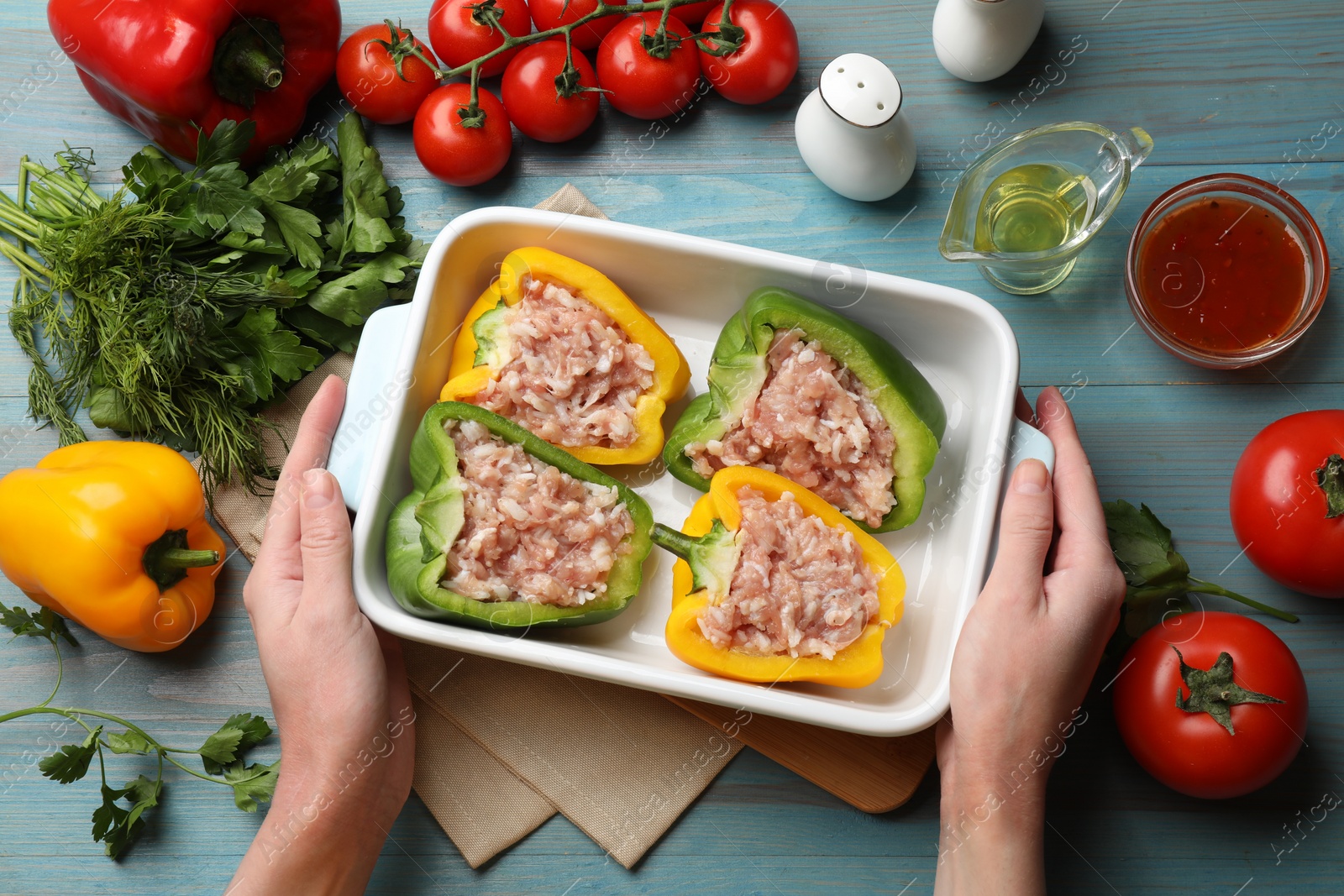 Photo of Woman holding dish with raw stuffed peppers at light blue wooden table, top view