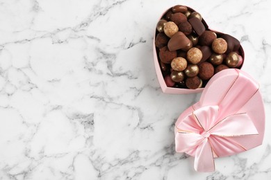 Different delicious chocolate candies in heart shaped box on white marble table, top view. Space for text