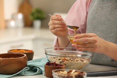 Photo of Making granola. Woman adding honey into bowl with mixture of oat flakes and other ingredients at table in kitchen, closeup