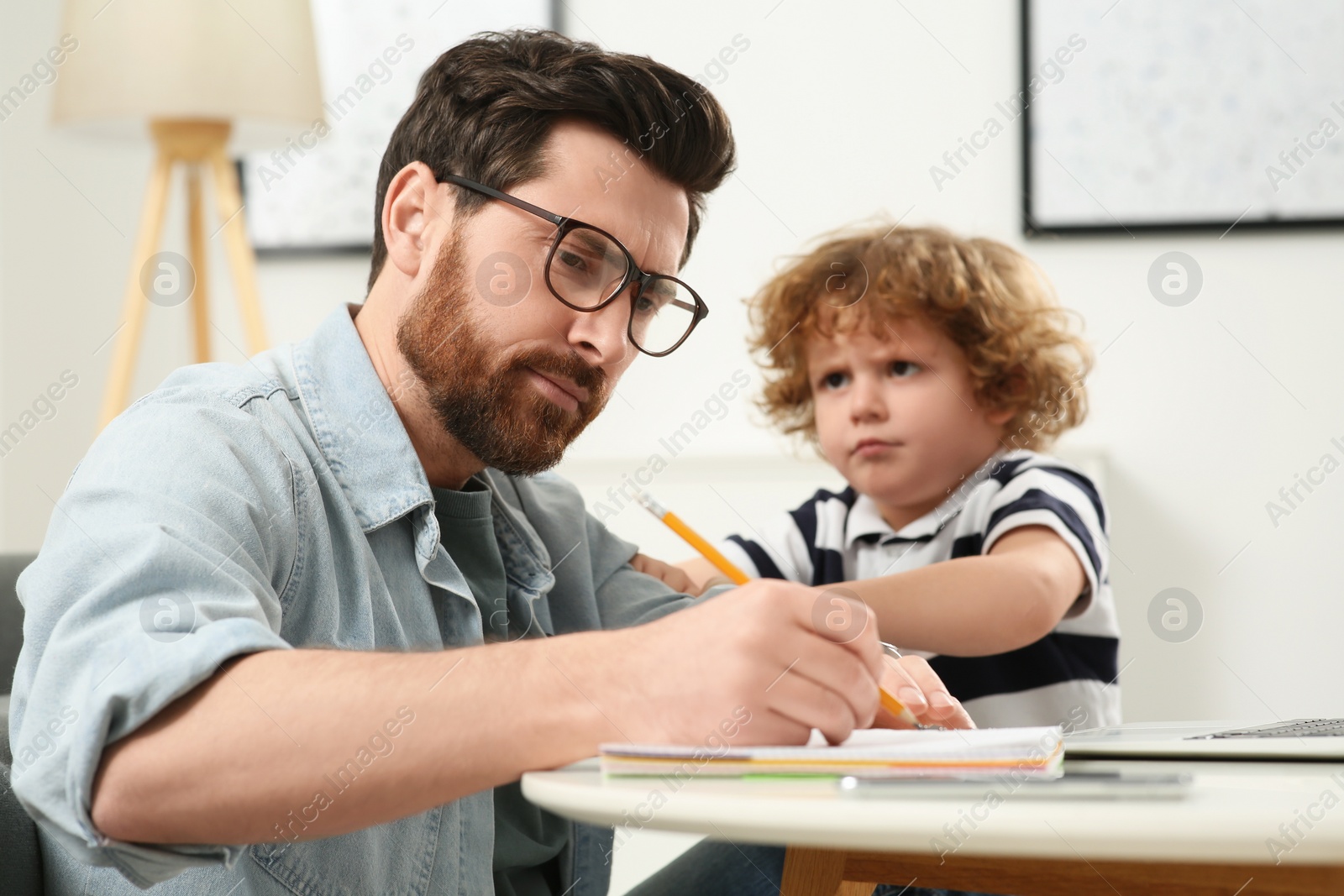 Photo of Little boy bothering his father at home. Man working remotely at desk