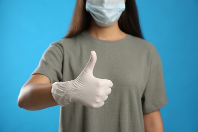Woman in protective face mask and medical gloves showing thumb up gesture on blue background, closeup