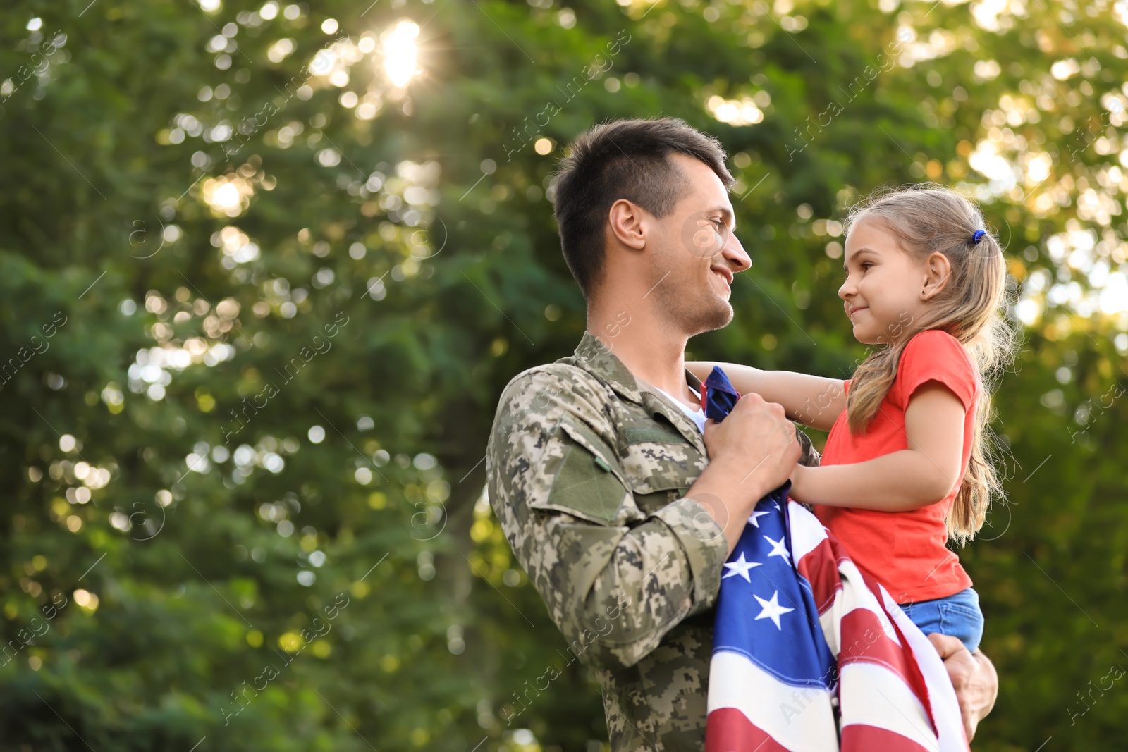 Photo of Father in military uniform with American flag holding his little daughter at green park