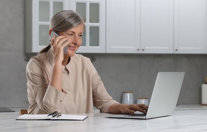 Photo of Beautiful senior woman talking on phone while using laptop at white marble table in kitchen