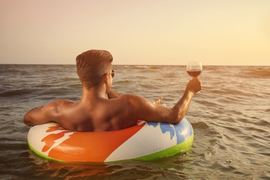 Photo of Man with glass of wine and inflatable ring resting in sea, back view