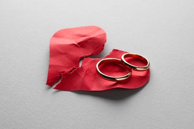Photo of Halves of torn red paper heart and wedding rings on white background. Broken heart