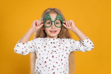 Photo of Smiling girl wearing decorative eyeglasses in shape of Christmas trees on orange background