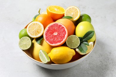 Photo of Different fresh citrus fruits and leaves in bowl on light table, closeup