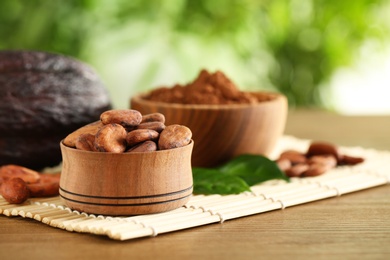 Photo of Wooden bowls of cocoa beans and powder near pod on table against blurred green background