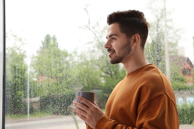 Photo of Happy handsome man with cup of coffee near window on rainy day