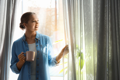 Photo of Woman with cup of coffee opening window curtain at home in morning