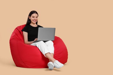 Happy woman with laptop sitting on beanbag chair against beige background, space for text