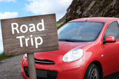 Image of Wooden plaque with inscription road trip. Car parked near mountains on cloudy day  