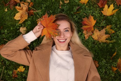 Photo of Smiling woman lying on grass and covering eye with autumn leaf, top view