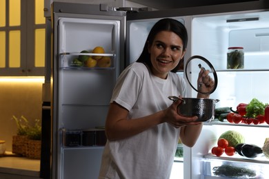 Young woman with pot near modern refrigerator in kitchen at night