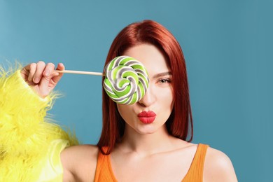 Photo of Stylish redhead woman covering eye with green lollipop on light blue background