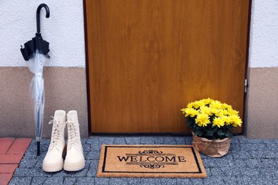Doormat with word Welcome, stylish boots, umbrella and beautiful flowers on floor near entrance
