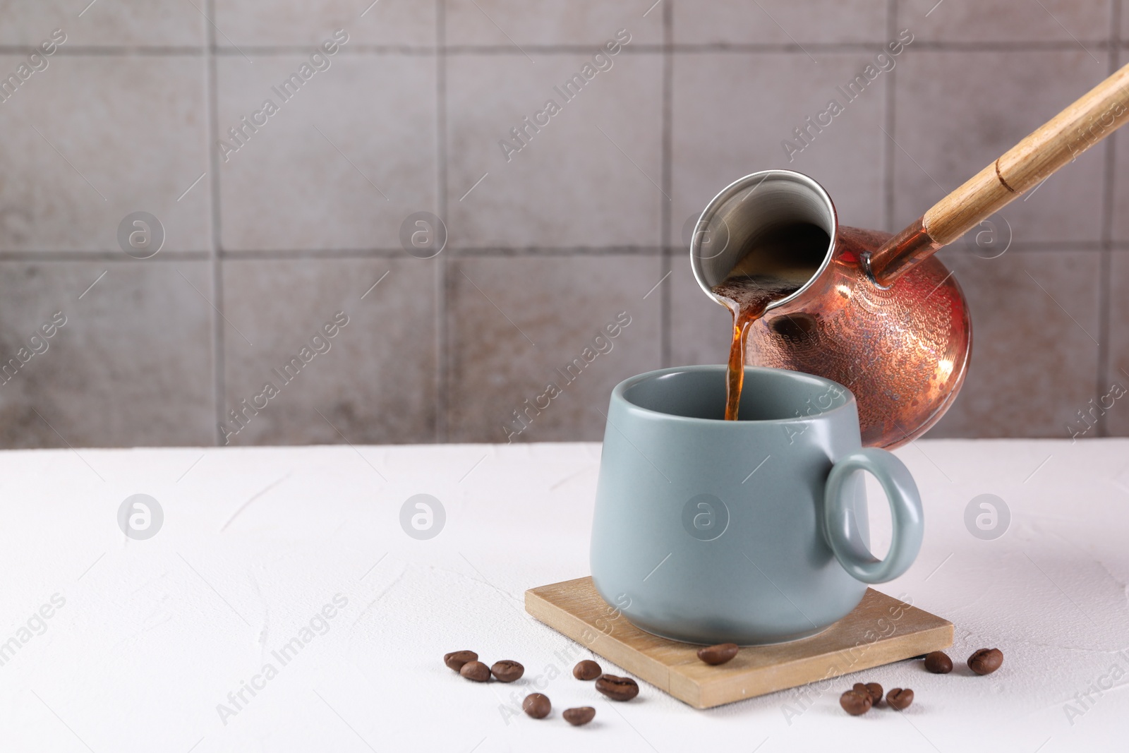 Photo of Pouring aromatic coffee from cezve into cup at white textured table, closeup. Space for text