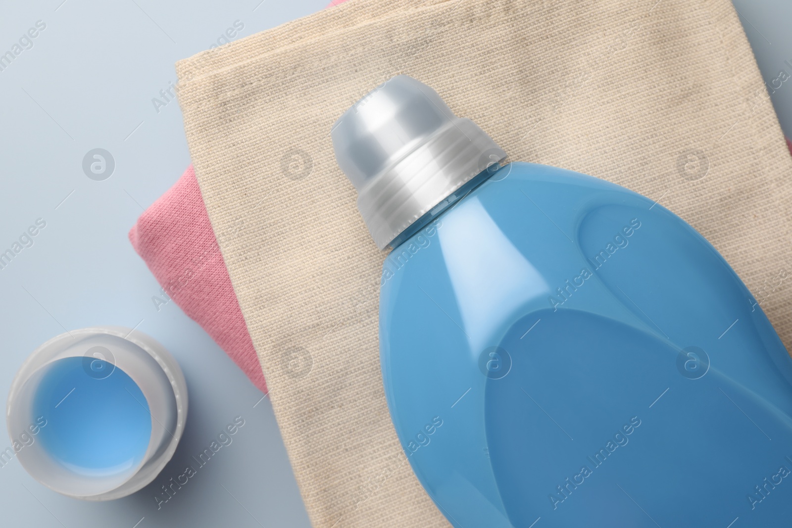 Photo of Bottle of fabric softener and stacked clean clothes on light blue background, flat lay