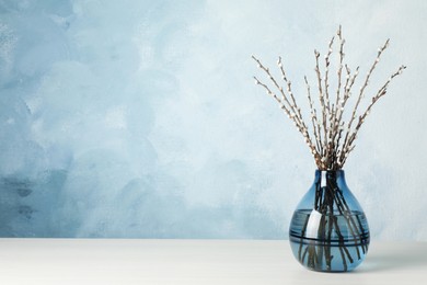 Beautiful pussy willow branches in glass vase on white wooden table against light blue background, space for text