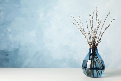 Photo of Beautiful pussy willow branches in glass vase on white wooden table against light blue background, space for text