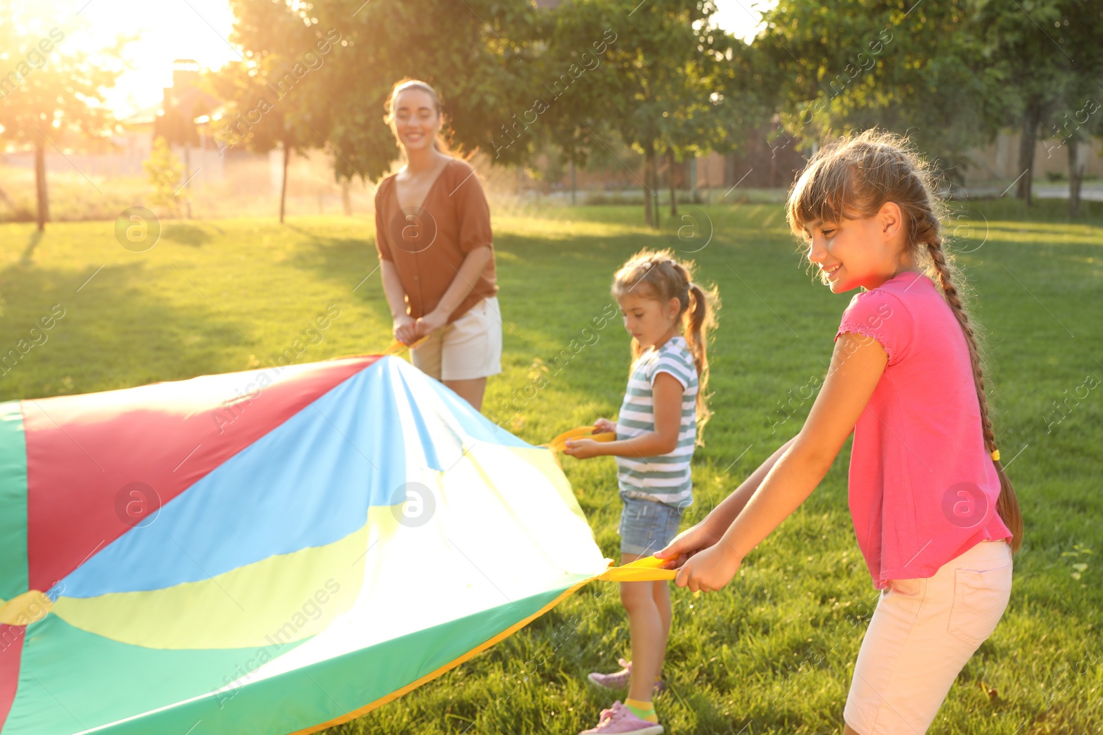 Photo of Group of children and teacher playing with rainbow playground parachute on green grass. Summer camp activity