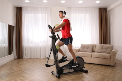 Photo of Man with headphones and towel using modern elliptical machine at home