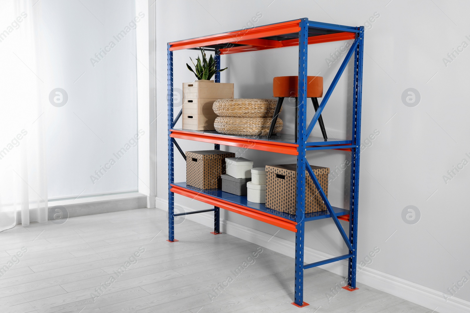 Photo of Metal shelving unit with different household stuff near light wall indoors