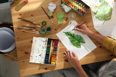 Photo of Young woman drawing leaf with watercolors at table indoors, top view
