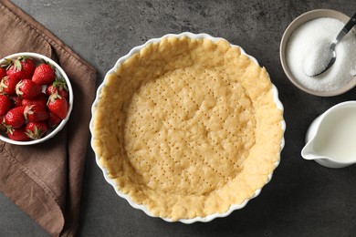 Making shortcrust pastry. Raw dough in baking dish, strawberries, sugar and milk on grey table, top view