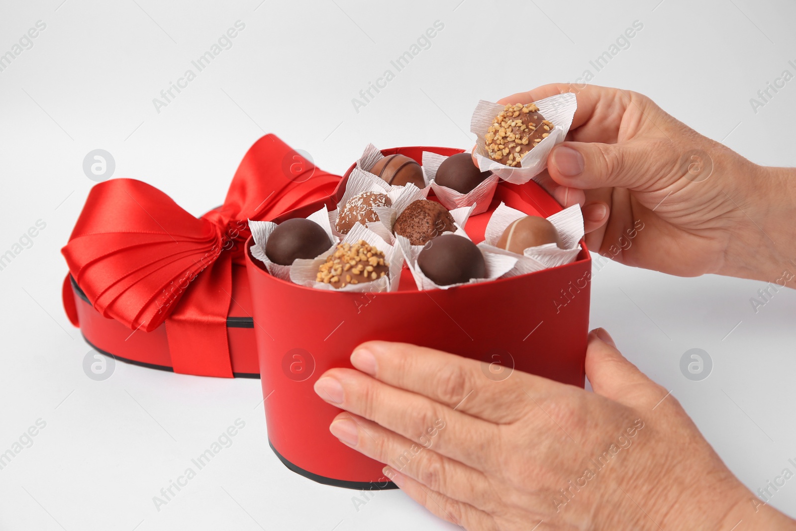 Photo of Woman taking delicious chocolate candy from heart shaped box on white background, closeup