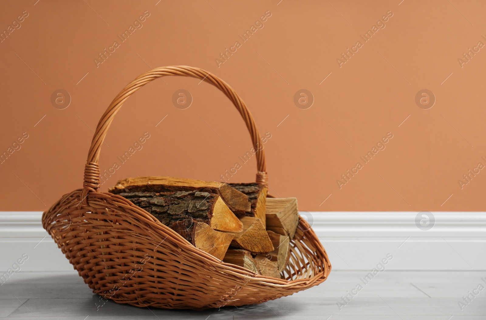 Photo of Wicker basket with firewood near brown wall indoors, space for text