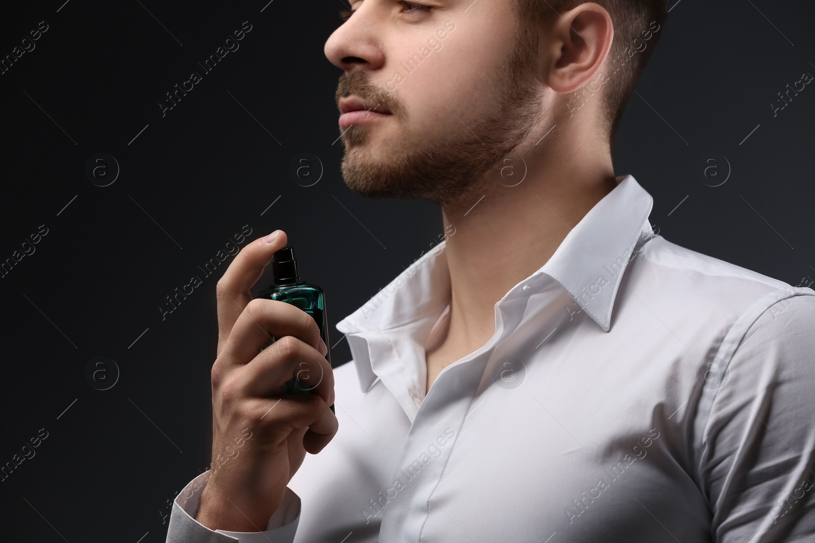 Photo of Handsome man in shirt using perfume on dark background, closeup