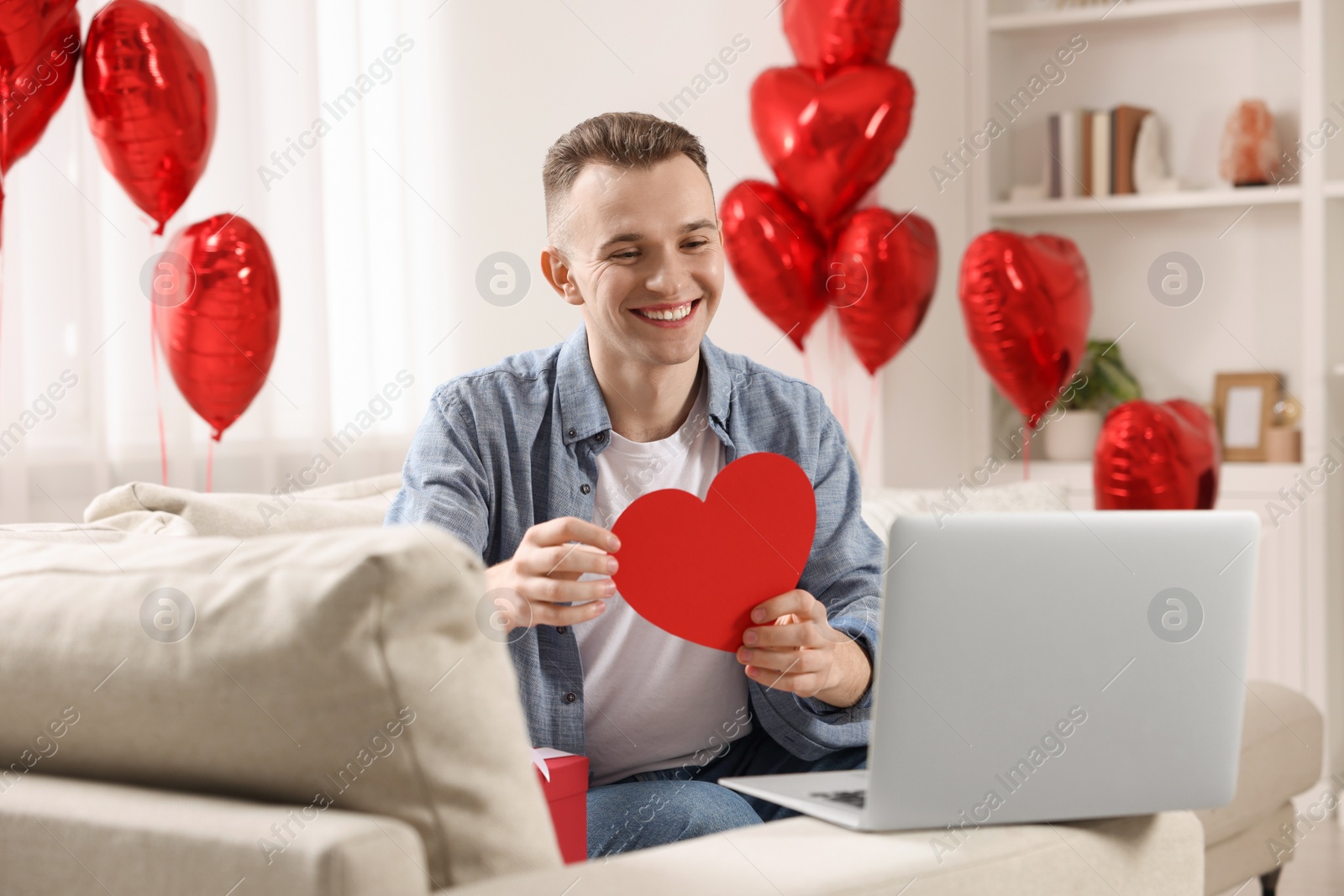 Photo of Valentine's day celebration in long distance relationship. Man holding red paper heart while having video chat with his girlfriend via laptop at home