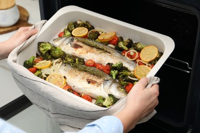 Photo of Woman taking baking dish with delicious fish and vegetables from oven in kitchen, closeup