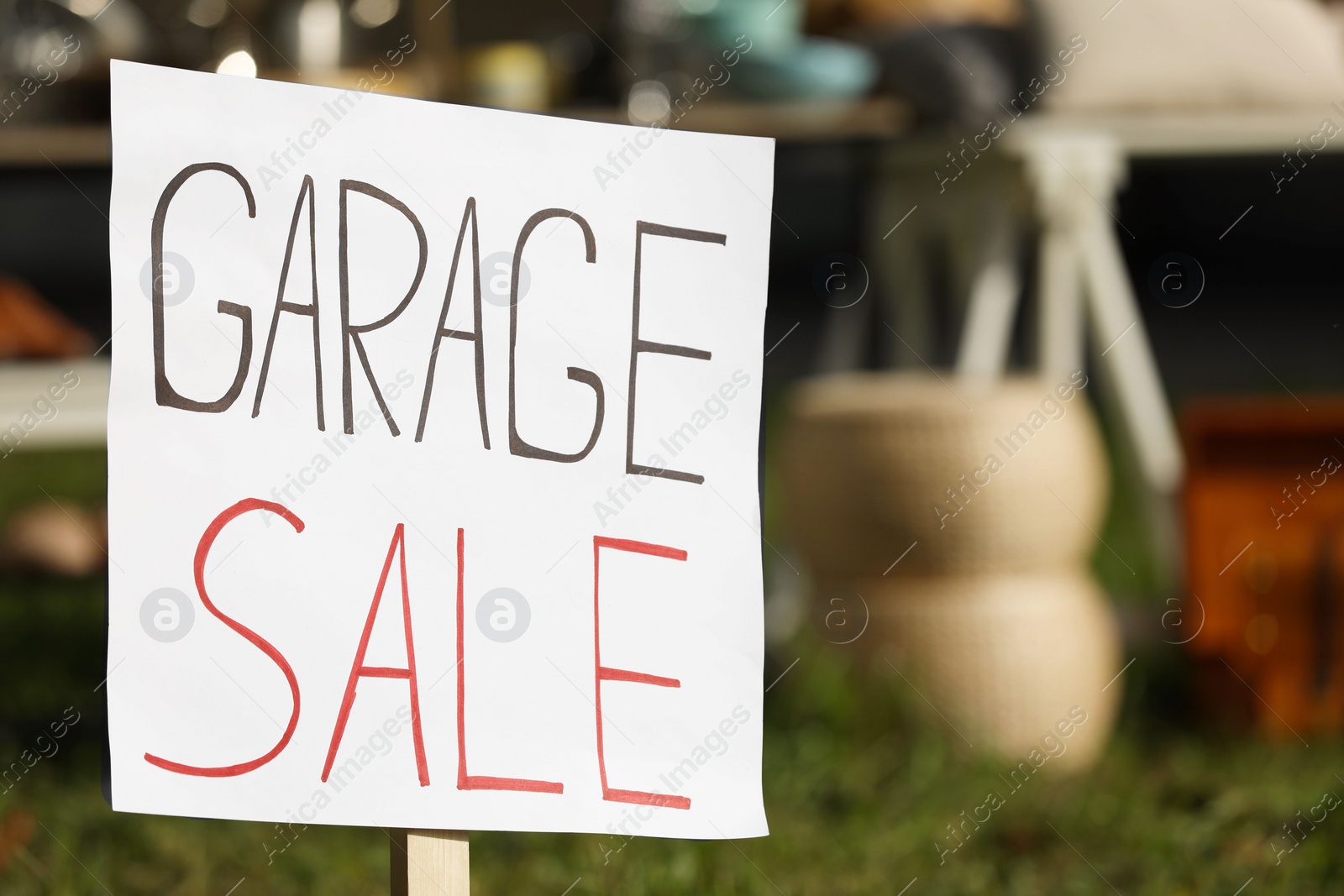 Photo of Sign Garage sale written on cardboard near tables with different stuff outdoors, closeup. Space for text